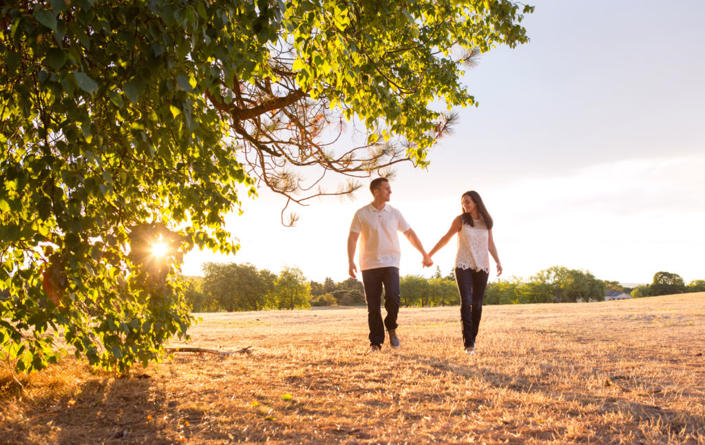 A man and woman are holding hands and walking in a grassy field during sunset at Discovery Park in Seattle, Washington. You can see green trees to the side where the sunlight is peeking in, plus a line of green trees in the background.