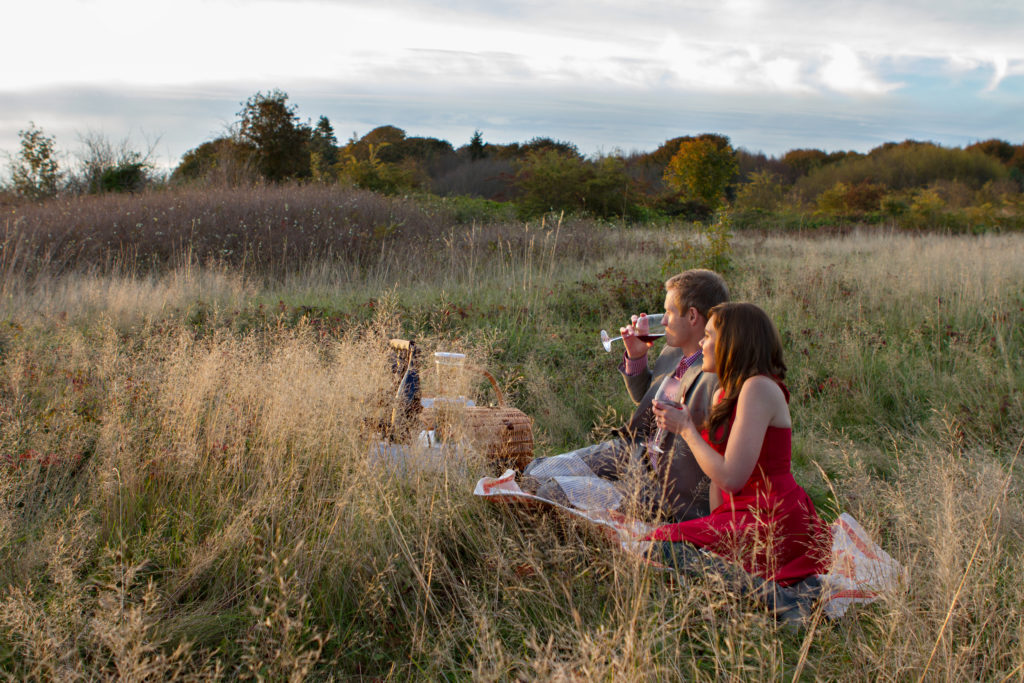 A man and woman are sitting on a picnic blanket in a grassy field during the golden hour sunset at Discovery Park in Seattle, Washington. They each have a wine glass in hand and are drinking while looking at the sunset. A picnic basket and food are set out in front of them. You can see a hillside with tall grass and green trees behind them.