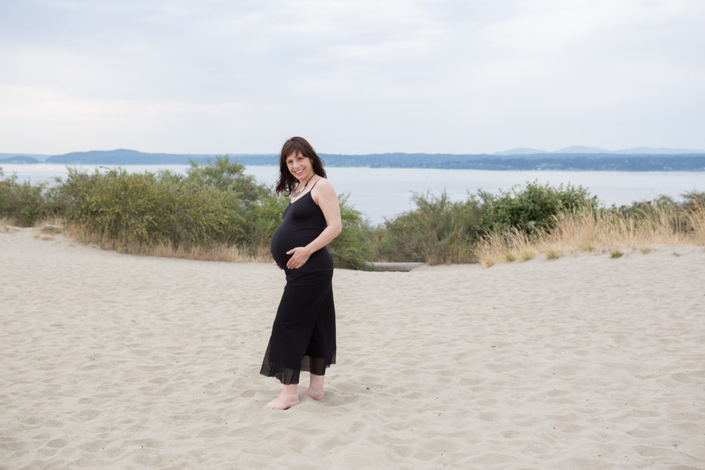 A pregnant woman is standing on the sandy bluff at Discovery Park in Seattle, Washington. Her hands are holding her stomach and she is smiling at the camera. You can see the Puget Sound in the background.