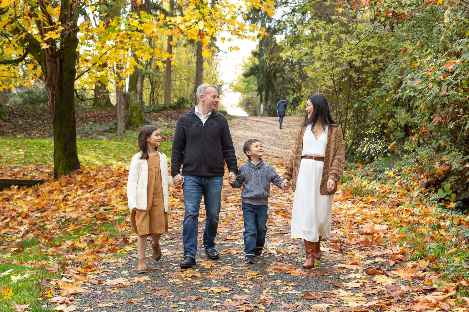 A family of four is walking together holding hands at Luther Burbank Park on Mercer Island near Seattle, WA. They're on a gravel path lined with trees and there are orange, yellow & red leaves on the ground.