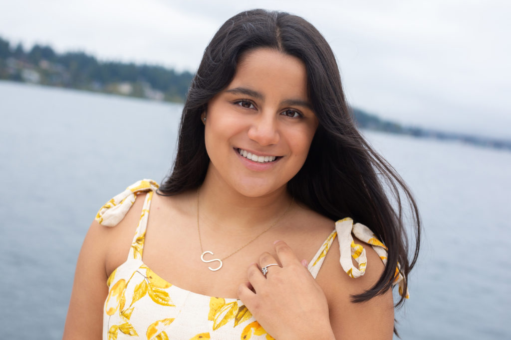 A high school senior girl is standing in front of Lake Washington at Luther Burbank Park on Mercer Island in Washington state. She is wearing a white sleeveless dress with yellow lemons on it. She is smiling & looking at the camera. The wind is blowing her hair.