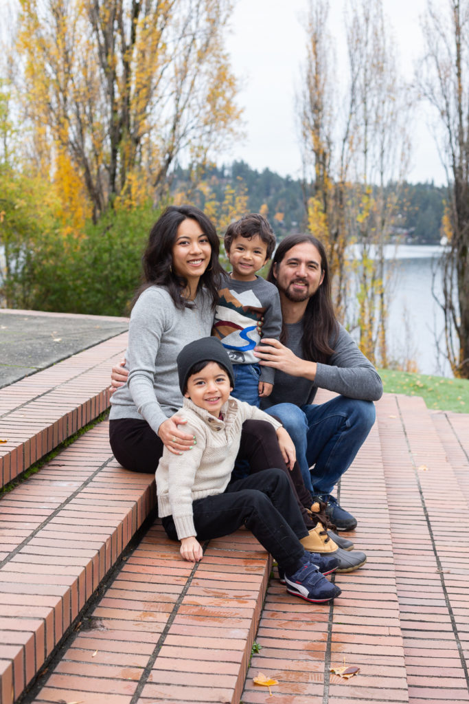 A mom, dad & their two small sons are sitting cuddled up together at Luther Burbank Park on Mercer Island near Seattle, WA in the fall. They're on brick steps with Lake Washington behind them.