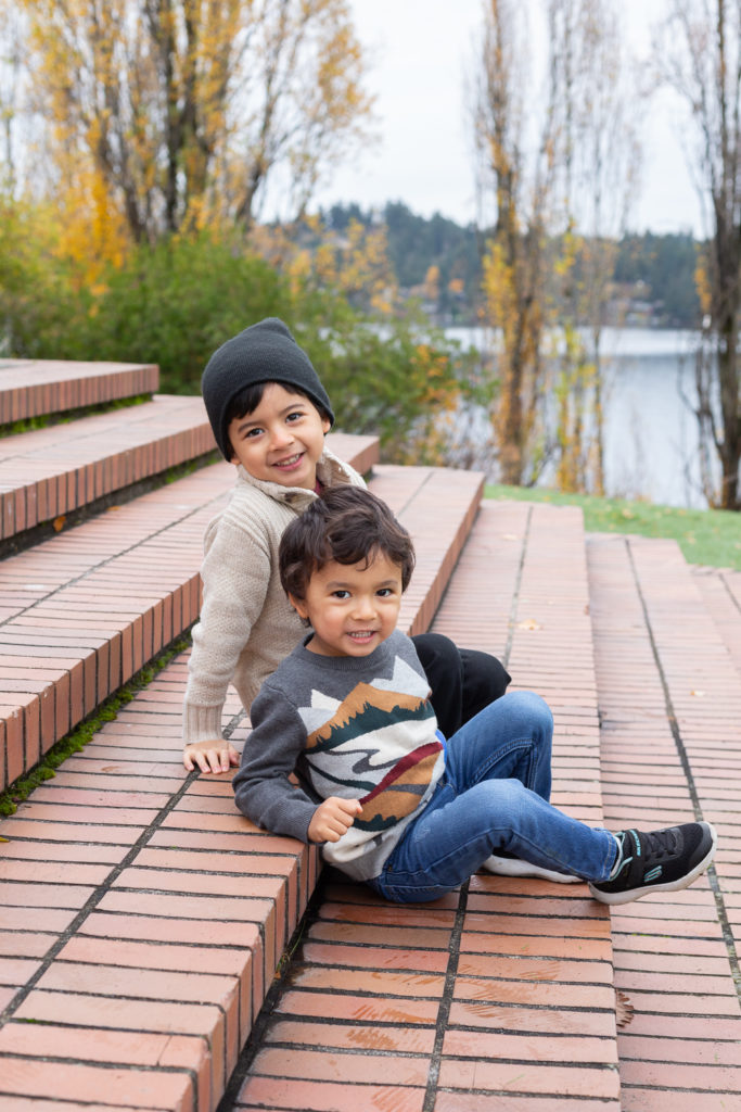 Two brothers, a toddler & little boy, are sitting cuddled up together at Luther Burbank Park on Mercer Island near Seattle, WA in the fall. They're sitting on brick steps and Lake Washington is behind them.