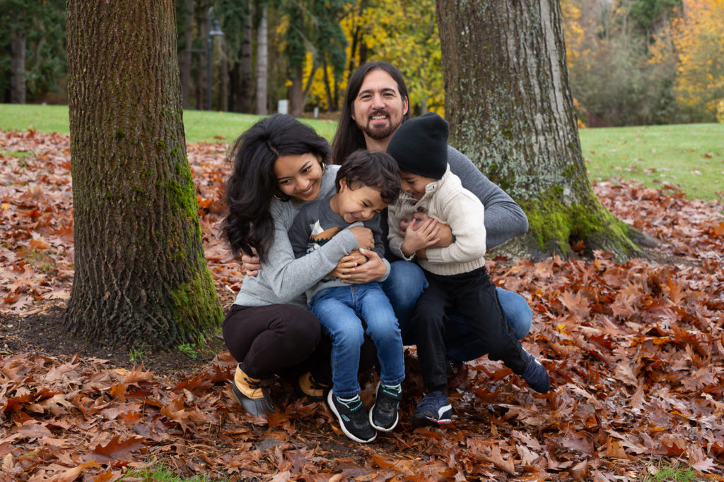 A mom & dad are tickling their two small sons while cuddled together at Luther Burbank Park on Mercer Island near Seattle, WA in the fall. Red & orange leaves are on the ground & there are trees in the background.