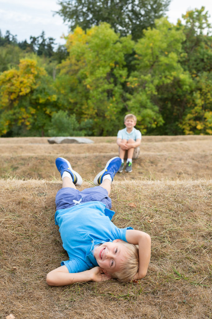 Two brothers, both elementary school aged, are posing for a portrait at Luther Burbank Park on Mercer Island near Seattle, WA in the summer. The younger brother is close to the camera and he's laying upside down on a hill, smiling at the camera. The older brother is sitting on the hill behind him and he's out of focus.