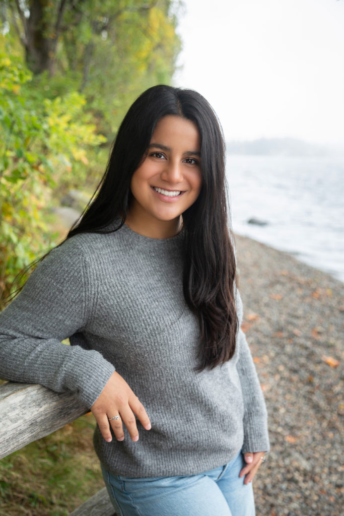 A high school senior girl is standing while leaning on a fence by the shoreline at Luther Burbank Park on Mercer Island in Washington state. There are green trees behind her and you can see some of Lake Washington next to her. She is wearing a gray sweater, blue jeans and black boots. She is smiling & looking at the camera.