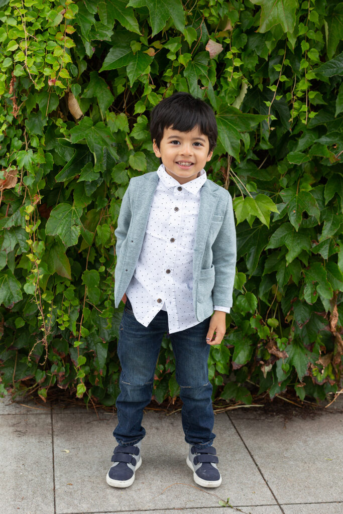 A small boy is standing by a column covered in green leaves at Heritage Park in Kirkland, Washington. He is wearing sneakers, jeans, a white shirt and a teal jacket. He has one hand in his pocket and is smiling at the camera. 