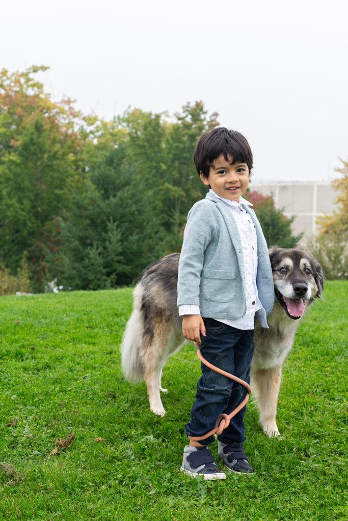 A small boy is standing on a grassy hill at Heritage Park in Kirkland, Washington. His dog is next to him and the dog's leash is wrapped around the boy's legs. The dog and the small boy are smiling at the camera. 