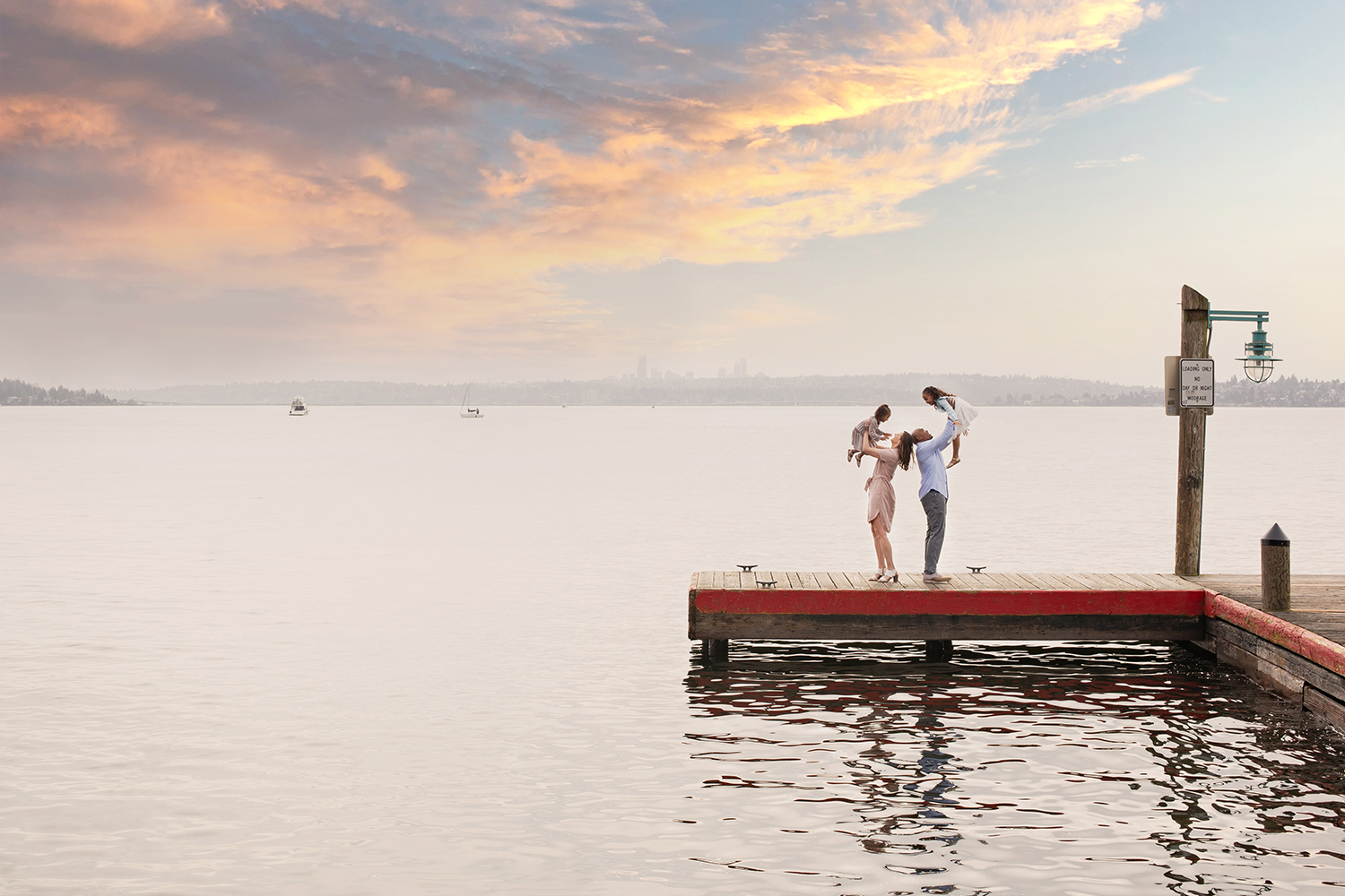 A family of four is standing together on a dock at Marina Park in Kirkland, Washington during sunset. The mom is holding her toddler daughter in the air while looking at her. The dad is holding the other daughter in the air & they're looking at each other.