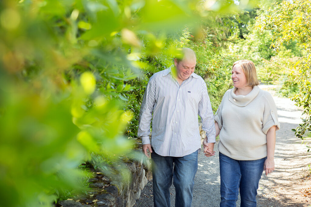 A man and woman are holding hands while laughing and walking along a path surrounded by green foliage at Heritage Hall in Kirkland, Washington. 