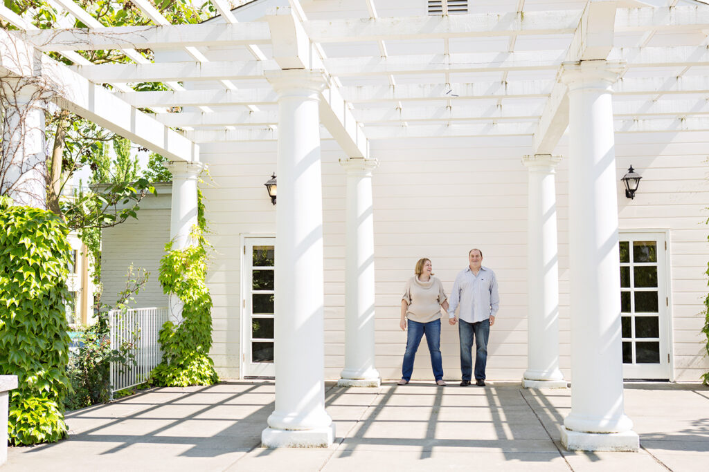A man and woman are holding hands and standing against a large white trellis and white wood building with columns at Heritage Hall in Kirkland, Washington. 