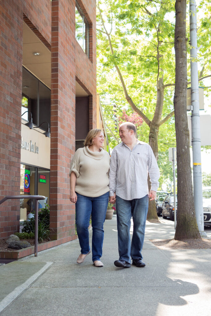 A man and woman are holding hands while laughing and walking along a city sidewalk surrounded by brick buildings and green trees in Kirkland, Washington. 