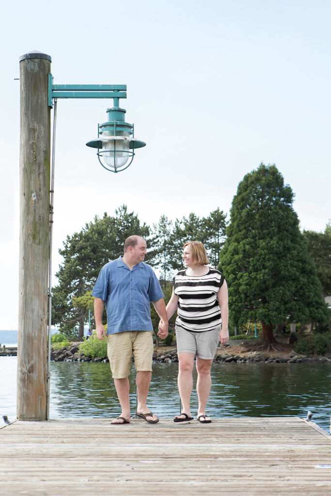 A man and woman are holding hands while laughing and walking on a dock at Marina Park in Kirkland, Washington. 