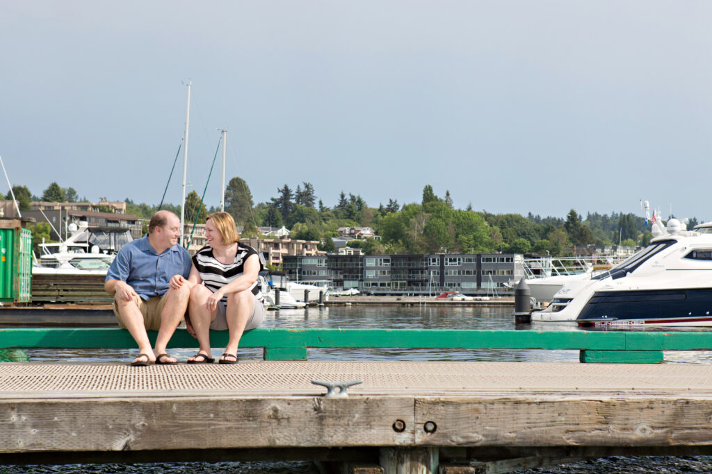 A man and woman are sitting next to each other and looking at one another on a dock at Marina Park in Kirkland, Washington. 
