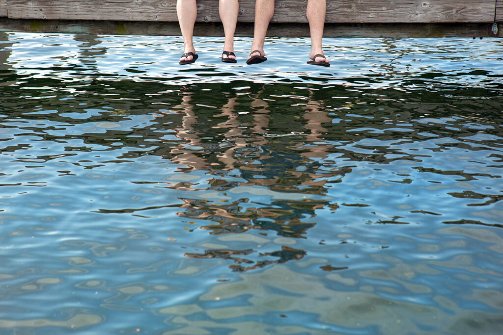 Two sets of legs dangle down from a dock and almost touch the water below.