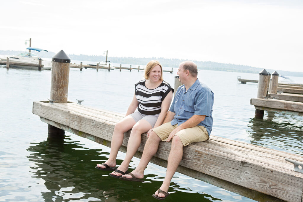 A man and woman are holding hands while laughing and sitting on a dock at Marina Park in Kirkland, Washington. 