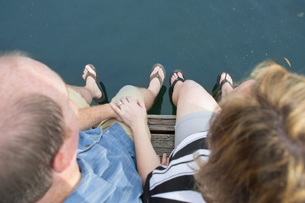 This image is taken from above. You see the top of a man's head and the top of a woman's head. The two are holding hands and there's an engagement ring on the woman's finger. They're sitting on a dock and their feet are almost touching the water below.