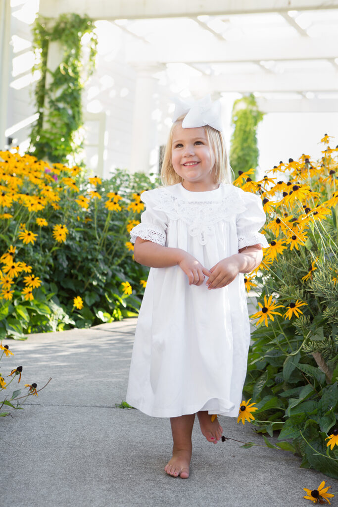 A toddler girl is standing in a pathway with vibrant yellow flowers surrounding her at Heritage Hall in Kirkland, Washington. She is wearing a long, white lace dress with bare feet and a big white bow in her blonde hair.