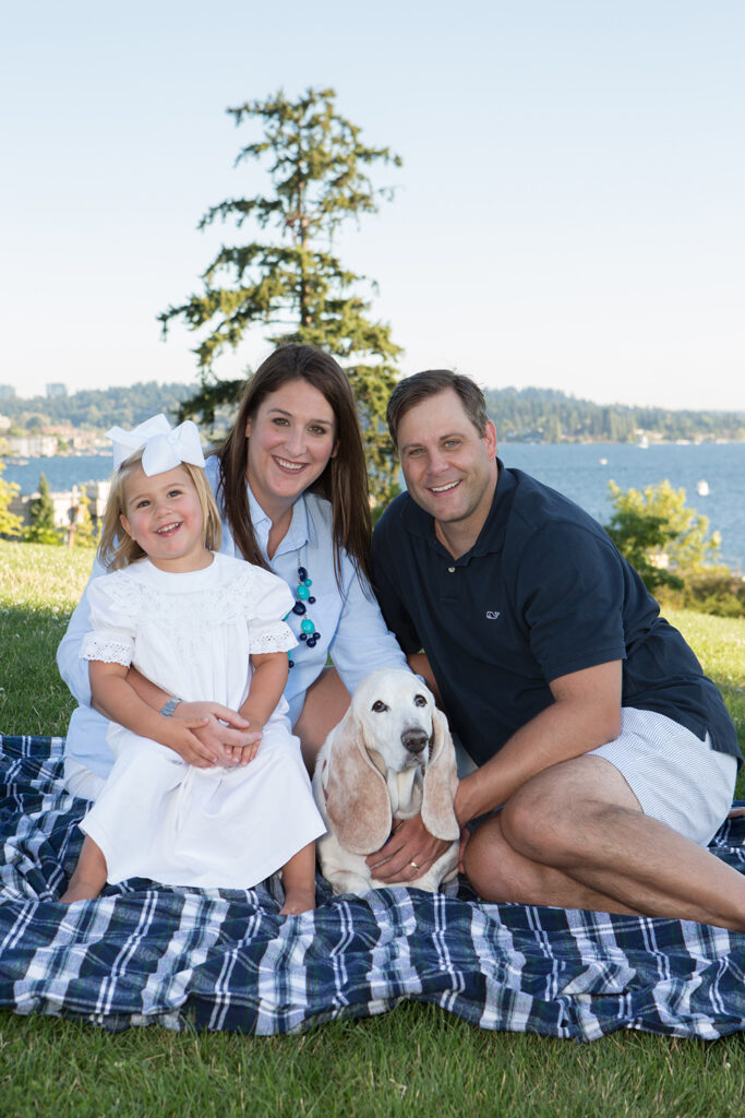 A mom, dad and toddler girl are sitting on a blanket with their basset hound in a grassy field overlooking Lake Washington at Heritage Park in Kirkland, Washington. The girl is sitting on her mom's lap and the family is cuddled up together. They're smiling and looking at the camera.