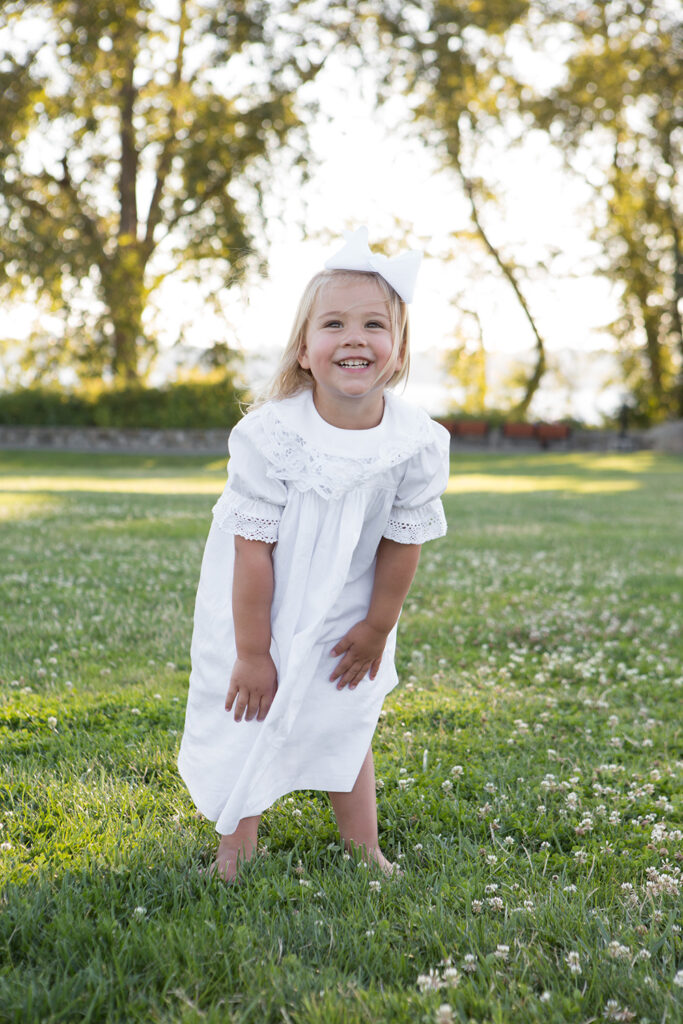 A toddler girl is standing in a grassy field with white clovers at Heritage Park in Kirkland, Washington. She is wearing a long, white lace dress with bare feet and a big white bow in her blonde hair.