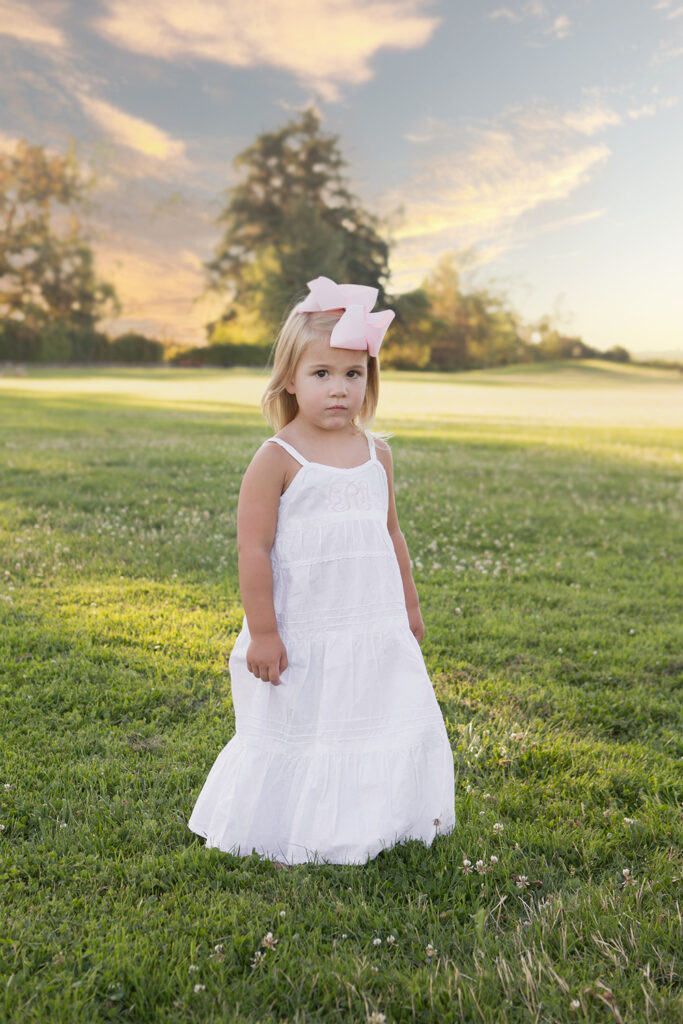 A toddler girl is standing in a grassy field with white clovers at Heritage Park in Kirkland, Washington at sunset. She is wearing a long, white lace dress with bare feet and a big pink bow in her blonde hair.