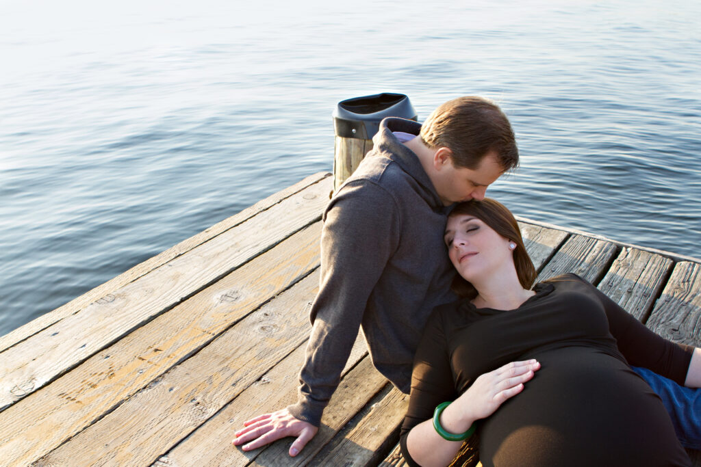 A pregnant woman and her husband are sitting on a dock at Marina Park in Kirkland, Washington. Lake Washington is visible behind them. The woman is laying her head on her husband's chest with her eyes closed. Her husband is kissing the top of her head. 