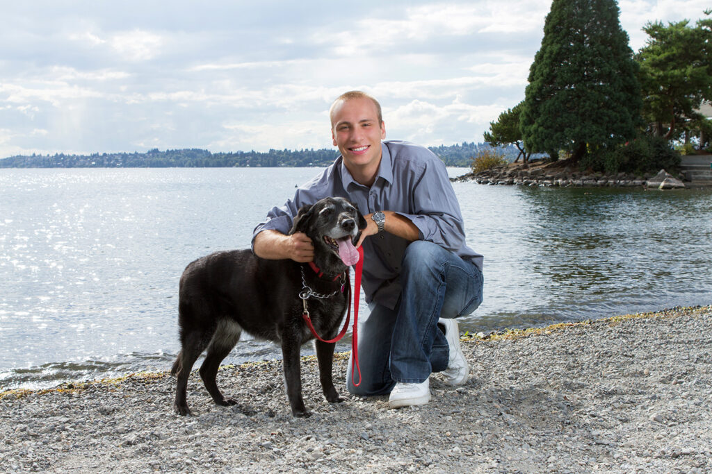 A high school senior boy is kneeing on the sand with his arm around his black dog at Marina Park in Kirkland, Washington. He is smiling at the camera and the dog has its tongue sticking out. Lake Washington and some trees are visible behind them.