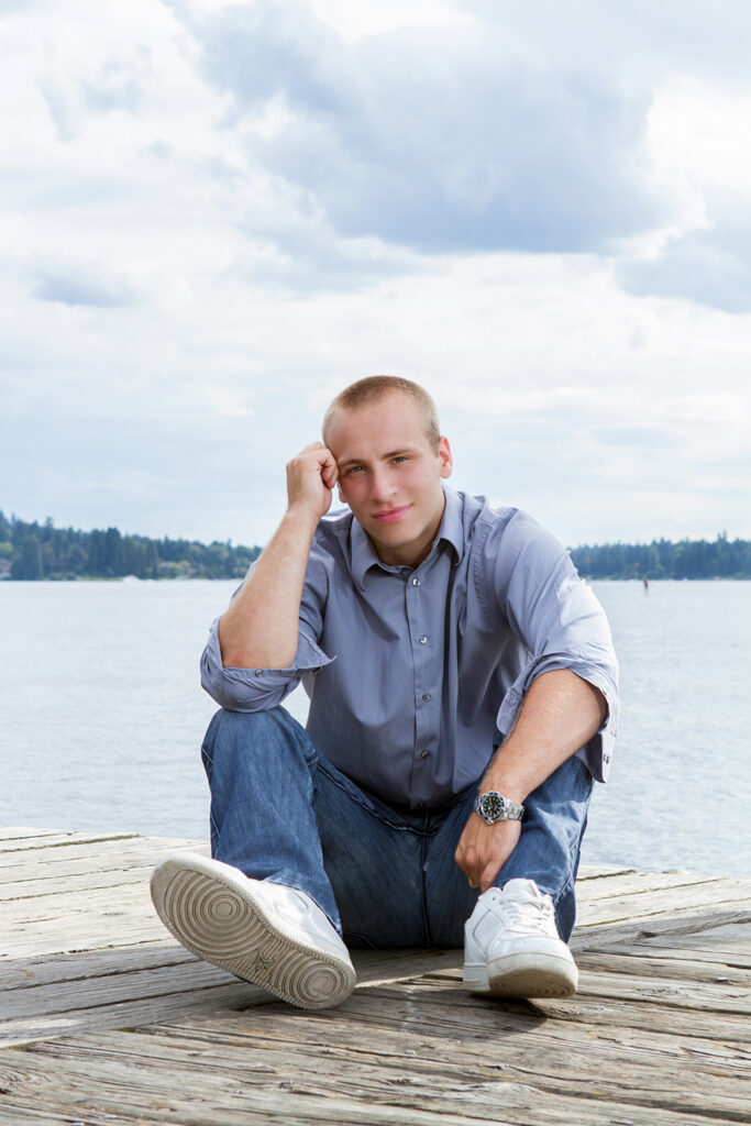 A high school senior boy is sitting on a dock at Marina Park in Kirkland, Washington. He has his head resting on his hand and is looking at the camera.